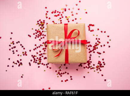 Geschenkbox in braun gefärbte Handwerk Papier gewickelt und mit roter Schleife auf Pastell rosa Hintergrund mit rotem Konfetti gebunden. Stockfoto