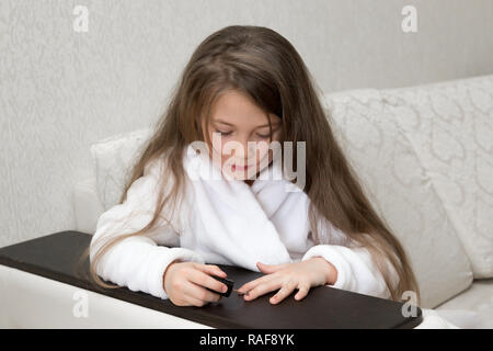 Das Mädchen von 3-4 Jahren in einem weißen Bademantel sitzt auf dem Sofa und Farben ihre Nägel mit Lack. Stockfoto