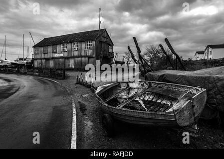 Schwarz & Weiß atmosphärischen Blick auf eine weggeworfene Boot am Straßenrand und ein Boot im Hintergrund beleuchten. Tollesbury Marina, Maldon, Essex, Großbritannien Stockfoto