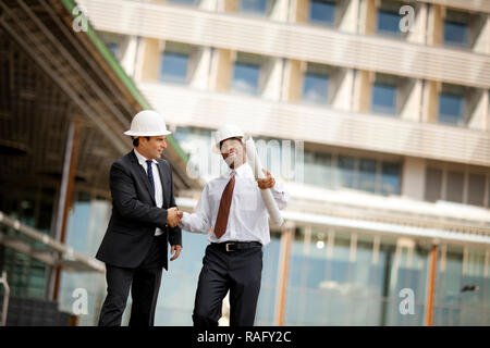 Zwei junge Ingenieure tragen Anzüge und hardhats zitternden Händen nach einer Diskussion über die Baupläne. Stockfoto