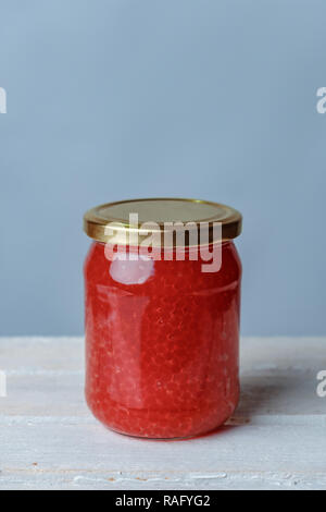 Ein großes Glas Glas mit rotem Kaviar stehen auf hölzernen Tisch und blauem Hintergrund gefüllt. Stockfoto