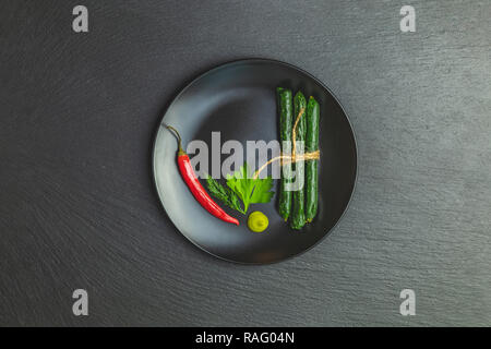 Kabanosy, Würstchen grün mit Wasabi von Schweinefleisch in der Platte auf einem schwarzen Stein Oberfläche mit Zugabe von frischen Kräutern und Gewürzen Stockfoto