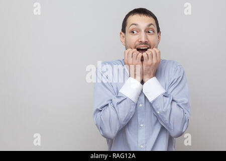 Portrait der nervösen stattlichen Borste Geschäftsmann in klassischem Hellblau shirt stehend, bitting seine Nägel und Wegsehen. indoor Studio shot, isolieren Stockfoto