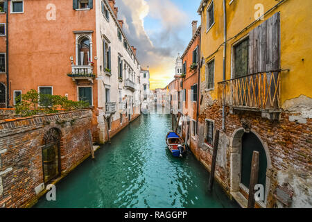 Ein kleines Boot Docks im smaragdgrünen Wasser von einem bunten Kanal in einem Wohngebiet, wie die Sonne unter Sturmwolken in Venedig, Italien. Stockfoto