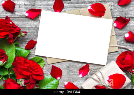 Valentines Tag Hintergrund. leere weiße Grußkarte mit rote Rose blumen blumenstrauss und Umschlag mit Blütenblättern und Geschenkbox mit rustikalen Holzmöbeln Hintergrund. Stockfoto