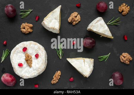 Zusammensetzung der Camembert oder Brie Käse mit Trauben, Walnüsse, Granatapfel und Rosmarin auf schwarzem Schiefer Hintergrund. Ansicht von oben. Gesundes Frühstück Stockfoto