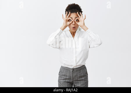 Charmante sorglos und kindliche weibliche Mitarbeiter in weißem Hemd und Hose Kreisen, mit der Hand über die Augen, Lippen, als ob Blick durch Fernglas auf etwas Unglaubliches und erstaunliches Stockfoto