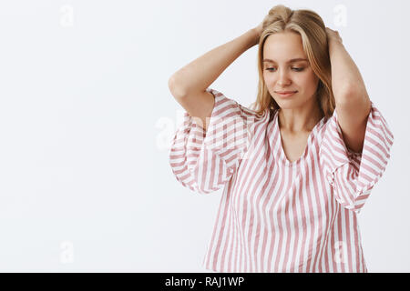 Taille - geschossen von Verträumten zart und feminin Blonde caucasian woman in pink gestreifte Bluse, Hände hinter dem Kopf, unten anstarren mit sanften leichtes Lächeln denken oder Imaging etwas Stockfoto