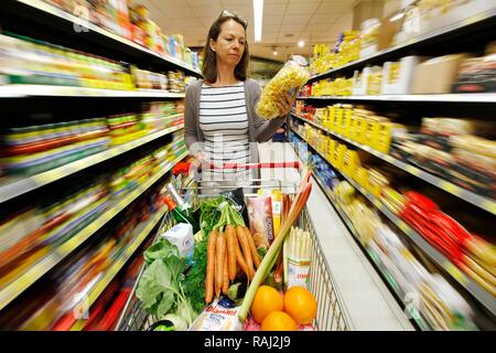 Frau treibt einen vollen Einkaufswagen durch einen Korridor der Lebensmittel Abschnitt beim Einkaufen in einem Self-service-Lebensmittelabteilung Stockfoto