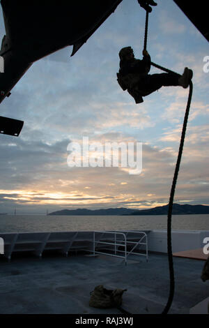 Ein Mitglied der Küstenwache Maritime Security Response Team West schnell - Seile aus einer 129 Rettung Flügel Hubschrauber auf das Deck der Hornblower Geist in einer Übung in der San Francisco Bay, 5. Dezember 2018. Teammitglieder sind in fortgeschrittene Techniken mit Bundes-, Landes- und lokalen Partnern ausgebildet, um die Nation aus einer Vielzahl von wasserbasierten Bedrohungen zu schützen. U.S. Coast Guard Foto von Chief Petty Officer John Masson. Stockfoto