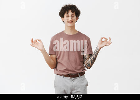 Bezaubernde curly-haired Kerl mit lustigen Schnurrbart im Lotussitz versuchen, Ruhe und Komfort Schließen der Augen und lächelnd mit ruhigen und entspannten Ausdruck Meditation über grauer Hintergrund Stockfoto