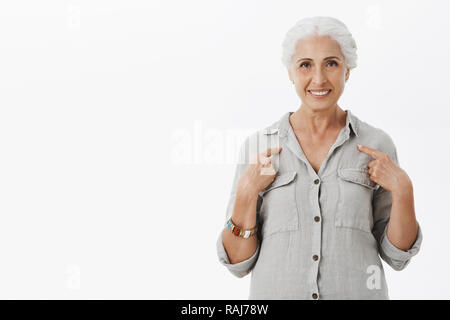 Studio shot von Art und nette ältere Mutter mit grauem Haar an sich zeigt mit zufrieden und selbstbewusst Lächeln was Ihr candidatute Gefühl zuversichtlich, dass sie keinen Job mehr als weiße Wand verarbeiten kann. Stockfoto