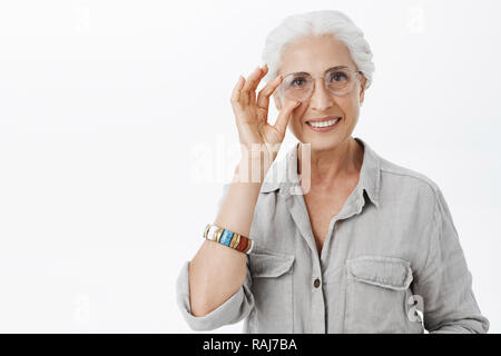 Taille-up Shot von charmanten Art und schönen Granny in Schaugläser berühren Rim von Brillen und freudig lächelnd hilft Enkel für Prüfungen freudige Posing vorbereiten und freuen uns über graue Wand Stockfoto