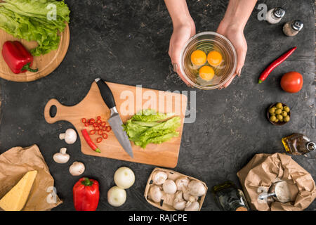 Teilweise mit Blick auf die Frau mit Schale mit Eier in der Nähe der Pizza Zutaten auf grauem Hintergrund Stockfoto