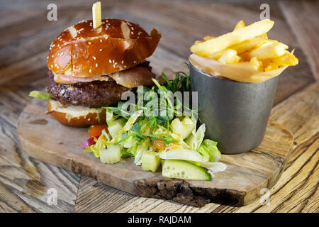 Burger serviert mit Pommes auf einem Holzbrett. Der Burger wird von Beilagensalat begleitet. Stockfoto