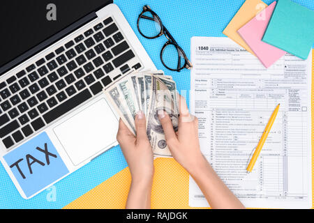 7/8-Ansicht von Frau mit Dollar Banknoten am Arbeitsplatz mit Laptop und Steuer Formular auf Hintergrund Stockfoto