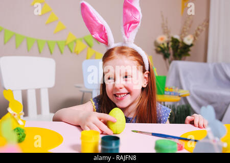 Fröhlich aktives Kind Mädchen mit roten Haaren und Rabbit Ears malt ein Ei mit gelben Farbe auf einem Hintergrund von Ostern Dekor und lächelt. Stockfoto