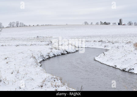 Es ist ein kleine Bucht oder Stream Wicklung entlang einer verschneiten Bauernhof Feld im Winter in einem ländlichen Gebiet mit landwirtschaftlichen Gebäuden im Hintergrund Stockfoto