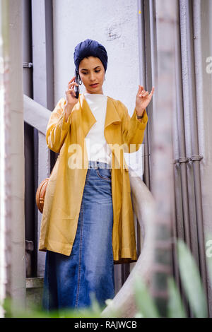 Eine junge und schöne muslimische asiatische Frau spricht auf Ihr Smartphone während des Tages. Sie trägt einen Turban Kopftuch aus religiösen Gründen. Stockfoto