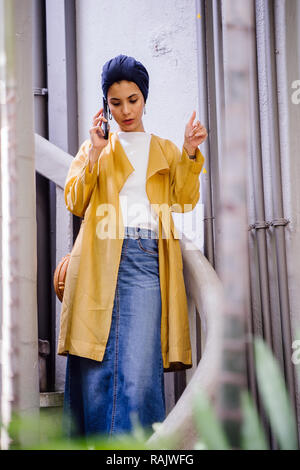 Eine junge und schöne muslimische asiatische Frau spricht auf Ihr Smartphone während des Tages. Sie trägt einen Turban Kopftuch aus religiösen Gründen. Stockfoto