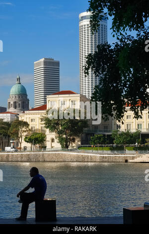 Ein Mann sitzt, der Singapore River in der Nähe von Boat Quay, im Hintergrund die Kaiserin, die Kuppel der National Gallery und Stamford Swissôtel Stockfoto