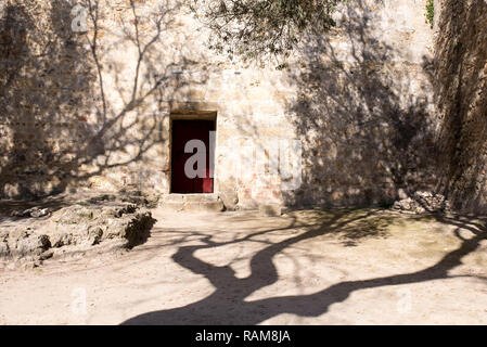 Eine rote Tür, wie der Eingang zu einer Festung in der Wüste Garten mit trockenen Böden und Olivenbäumen. Stockfoto