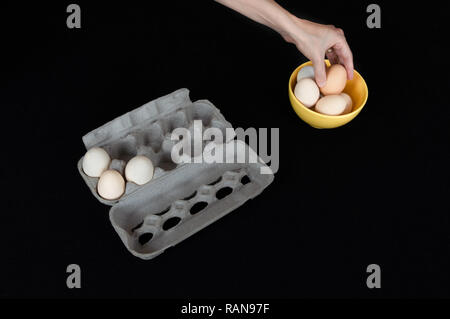 Weibliche Hand ein Ei aus gelben Schüssel. Karton ei Box mit drei Eier auf schwarze Hintergrund. Stockfoto