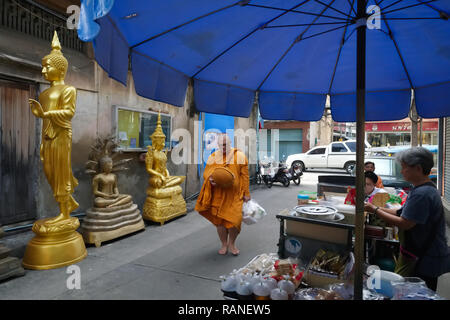 Ein Mönch auf seinen Morgen Almosen Runde in Bamrung Muang Road, Bangkok, Thailand, vorbei Buddha Figuren außerhalb eines Buddha, Fabrik und eine Garküche Stockfoto