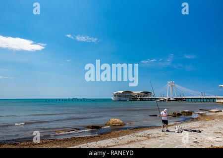 DURRES, Albanien - 17. MAI 2017: Junge Fischer legt seine Angelausrüstung auf den Strand der Stadt im sonnigen blauen Himmel Frühlingstag. Die moderne Architektur Ve Stockfoto