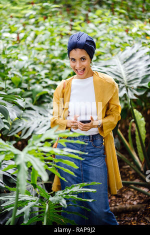 Porträt einer jungen und attraktiven Malay asiatische Muslimin in einem turban hijab Kopftuch stehend in einem Park und lächelnd, als Sie Ihr Smartphone verwendet. Stockfoto