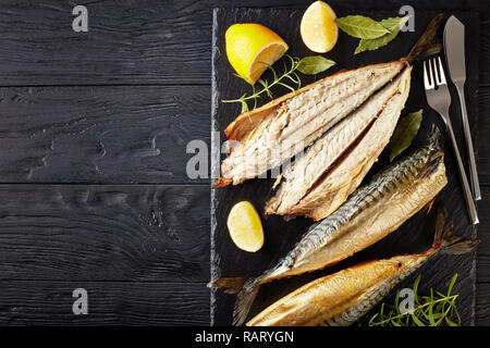 Nahaufnahme der geräucherte Makrele, serviert auf einem schwarzem Schiefer Fach mit Salz, Pfeffer, Rosmarin und Zitronenscheiben, silber Gabel und Messer, Ansicht von oben, Fla Stockfoto