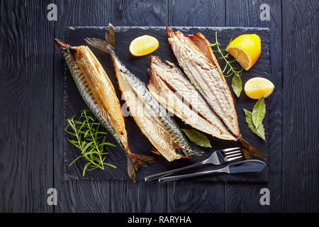 Nahaufnahme der geräucherte Makrele, serviert auf einem schwarzem Schiefer Fach mit Salz, Pfeffer, Rosmarin und Zitronenscheiben, Messer und Gabel, Ansicht von oben Stockfoto