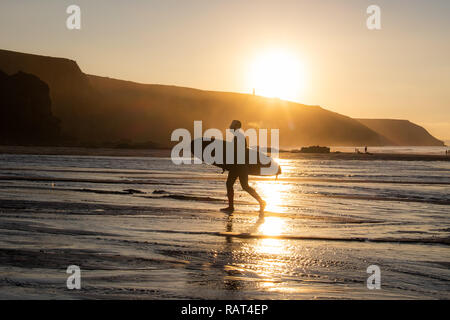 Single surfer zurück lit durch den Sonnenuntergang, seinem Surfbrett, weg von den Wellen und der Strand am Ende des Tages bei Porthtowan, Cornwall. Stockfoto