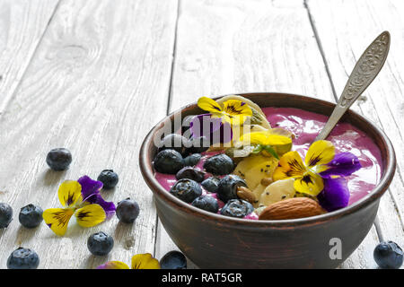 Smoothie Schale mit einem Löffel garniert mit frischen Beeren, Banane, Chia Samen, Nüsse und Blumen für eine gesunde vegetarische Ernährung Frühstück. Nach oben Schließen Stockfoto