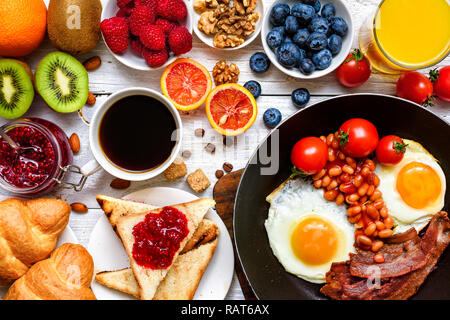 Frühstück mit Kaffee, Spiegeleier, Speck, Bohnen, Toast, Croissants, Orangensaft mit Früchten und Beeren. top View Stockfoto