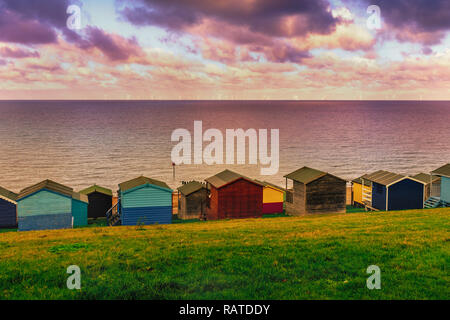 Moody wetter Patches erstellen die Sonne zwischen den Wolken am Meer in Tankerton, Whitstable, Kent. Reihen von Holz- Strand Hütten säumen die Stockfoto