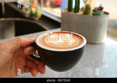 Eines Mannes Hand eine Tasse Cappuccino mit einzigartigen Latte Art, unscharfe Tabelle im Hintergrund Stockfoto