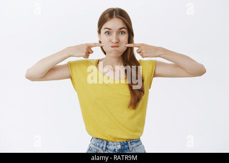 Charismatische lustig und fröhlich charmante junge Frau im gelben T-Shirt schmollend stossen Wangen mit Index Finger und lächelte, als wenn Atem spielerisch um Narren auf weißem Hintergrund Stockfoto