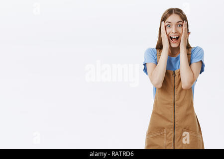 Verstand verrückt von Awesome überraschen. Portrait von beeindruckt und erstaunt, hübsche junge Frau in süßen Overalls mit Palmen zu Wangen breit grinsend mit Staunen und Freude über graue Wand Stockfoto