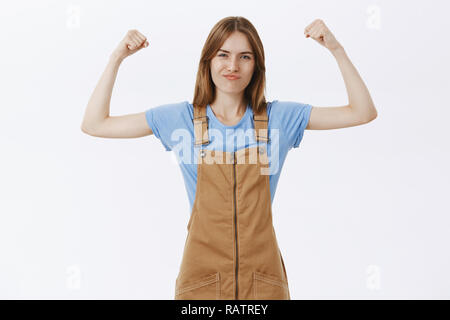 Portrait von stolzen, selbstbewussten und starken weiblichen femenist im stilvollen Overalls über blaues T-Shirt heben die Hände zeigen Muskeln und Bizeps grinste Selbstbewusst heben das Selbstwertgefühl tun Fitness Übungen Stockfoto