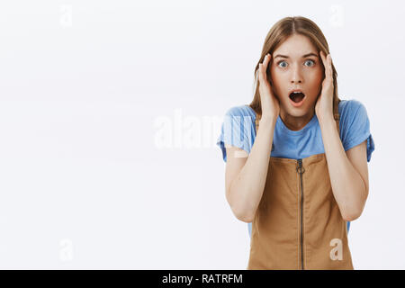 Taille - geschossen von geschockt, sprachlos und fassungslos attraktive junge Frau in braunen Overalls holding Palmen am Tempel drop Kiefer von Schock und Staunen stehen fassungslos und bestürzt über graue Wand Stockfoto