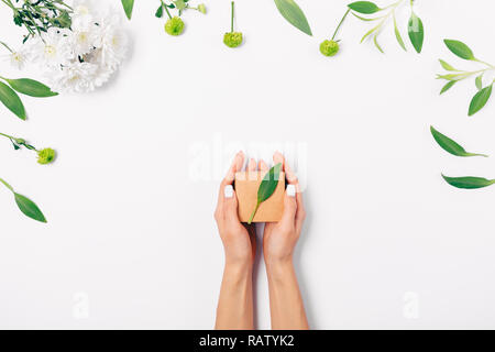 Blick von oben Frau Hände halten kleine Geschenkbox braun eco Papier, flach Komposition von frischen Blumen und grüne Blätter auf weißem Hintergrund. Stockfoto