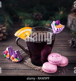 Dekorativer Löffel mit Kuchen im Glas mit Tee und orangen Scheibe in der Nähe von Rosa macarons auf hölzernen Hintergrund zur Weihnachtszeit Stockfoto