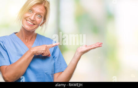 Blonde Frau mittleren Alters tragen Arzt Krankenschwester uniform über isolierte Hintergrund erstaunt und lächelt in die Kamera während der Präsentation und mit der Hand zeigen Stockfoto