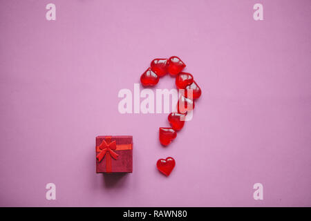 Eine Box mit einem Geschenk und einer Reihe von Fragezeichen aus kleinen roten Herzen gelegt. Konzept für den Valentinstag oder eine andere Liebe. Ein Fragezeichen bedeutet eine Überraschung. Stockfoto