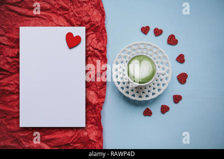 Eine Tasse mit einem leckeren und aromatischen morgen Matcha Latte Kaffee auf einer festlichen Hintergrund mit vielen Herzen. Neben dem roten Hintergrund ist ein weisses Blatt für Text. Feier der Valentinstag oder Tag der Frau oder andere Liebe. Stockfoto