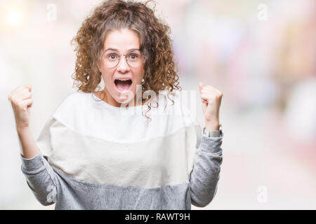 Schöne Brünette lockige Haare junge Mädchen Tragen einer Brille über isolierte Hintergrund feiern überrascht und für den Erfolg mit erhobenen Armen und Op erstaunt Stockfoto