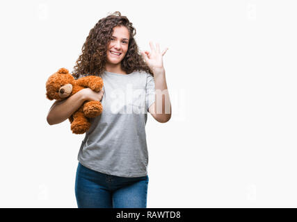 Junge brünette Mädchen, dass Teddybären über isolierte Hintergrund tun ok Zeichen mit den Fingern, ausgezeichnete Symbol Stockfoto