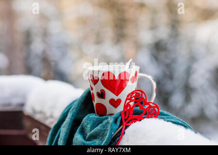 Heiße Schokolade oder Kaffee, Rotes Herz in der Nähe der Schale, Winter Hintergrund mit Lichter aus. Winter oder Valentinstag Hintergrund. Grußkarte. Weiße Tasse heißes Getränk und rotem Filz Herzen. Platz kopieren Stockfoto
