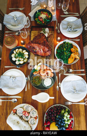 Moderne australische Weihnachtsessen Tabelle mit glasierten Schinken, Garnelen, Kartoffeln und Dill, asiatische Grüns, Christmas Pudding, Hackfleisch Obstkuchen bei Kerzenschein. Stockfoto